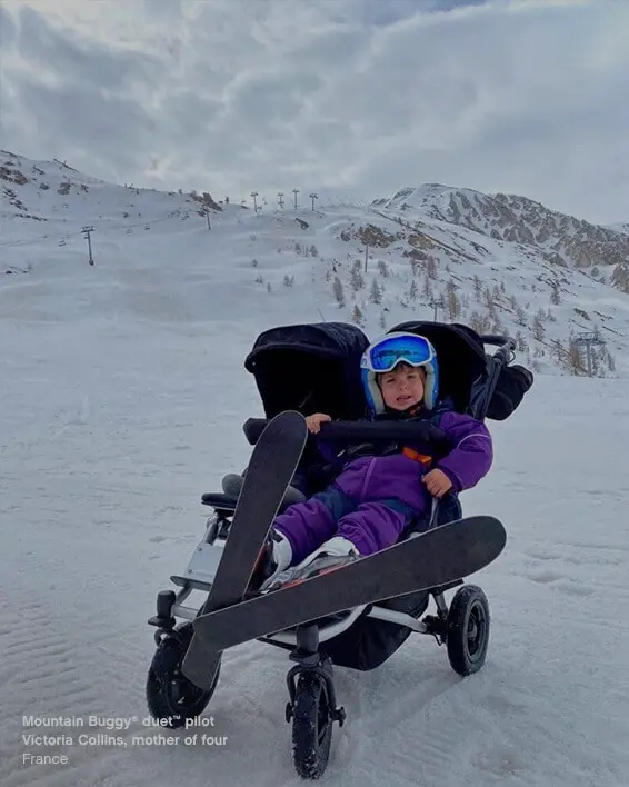 child dressed in ski suit with both skis on sitting in duet™ four wheeled double buggy at skiing field - Mountain Buggy life without limit